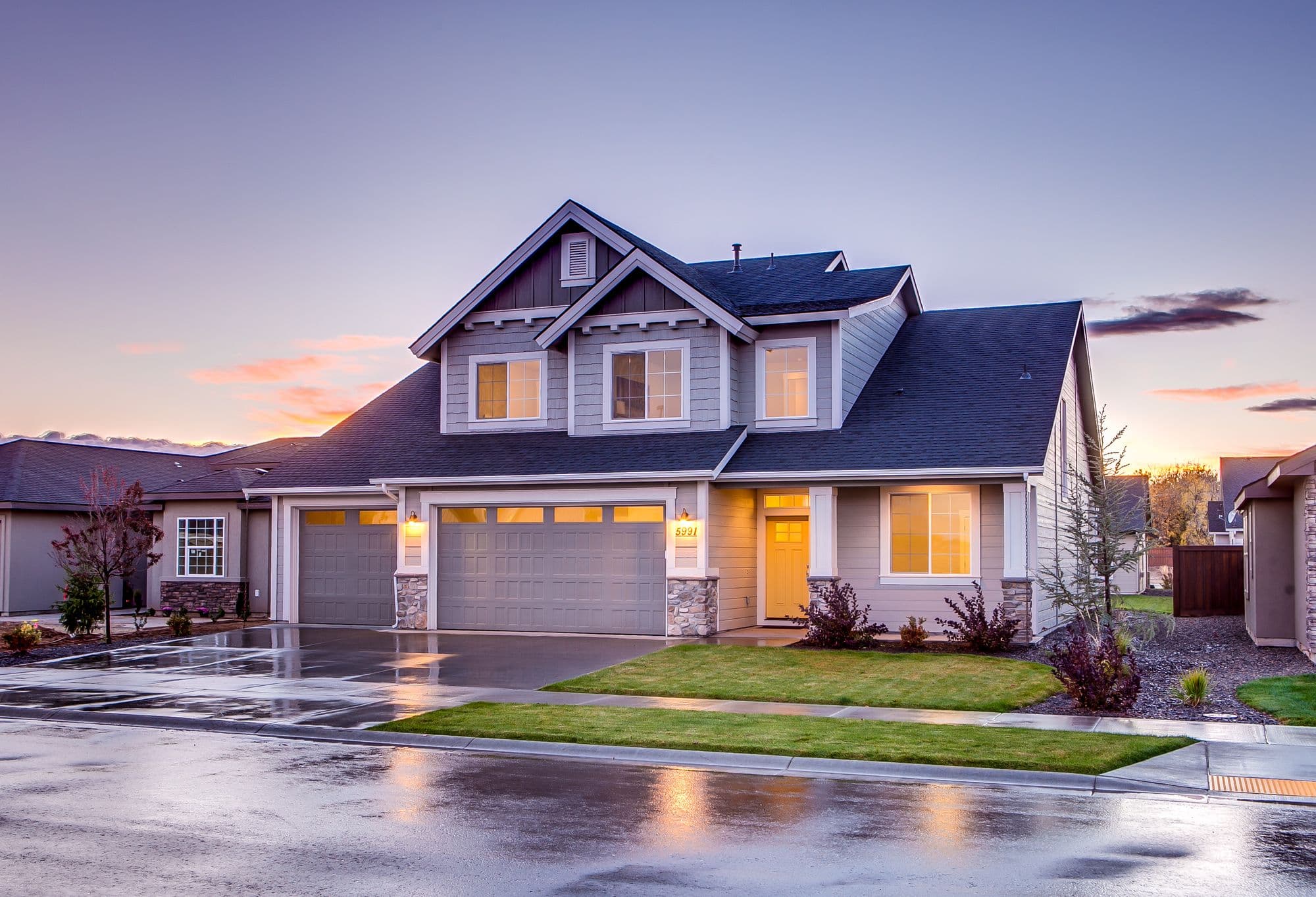 Two storey suburban house at dusk with a three car garage, lit windows and a front lawn