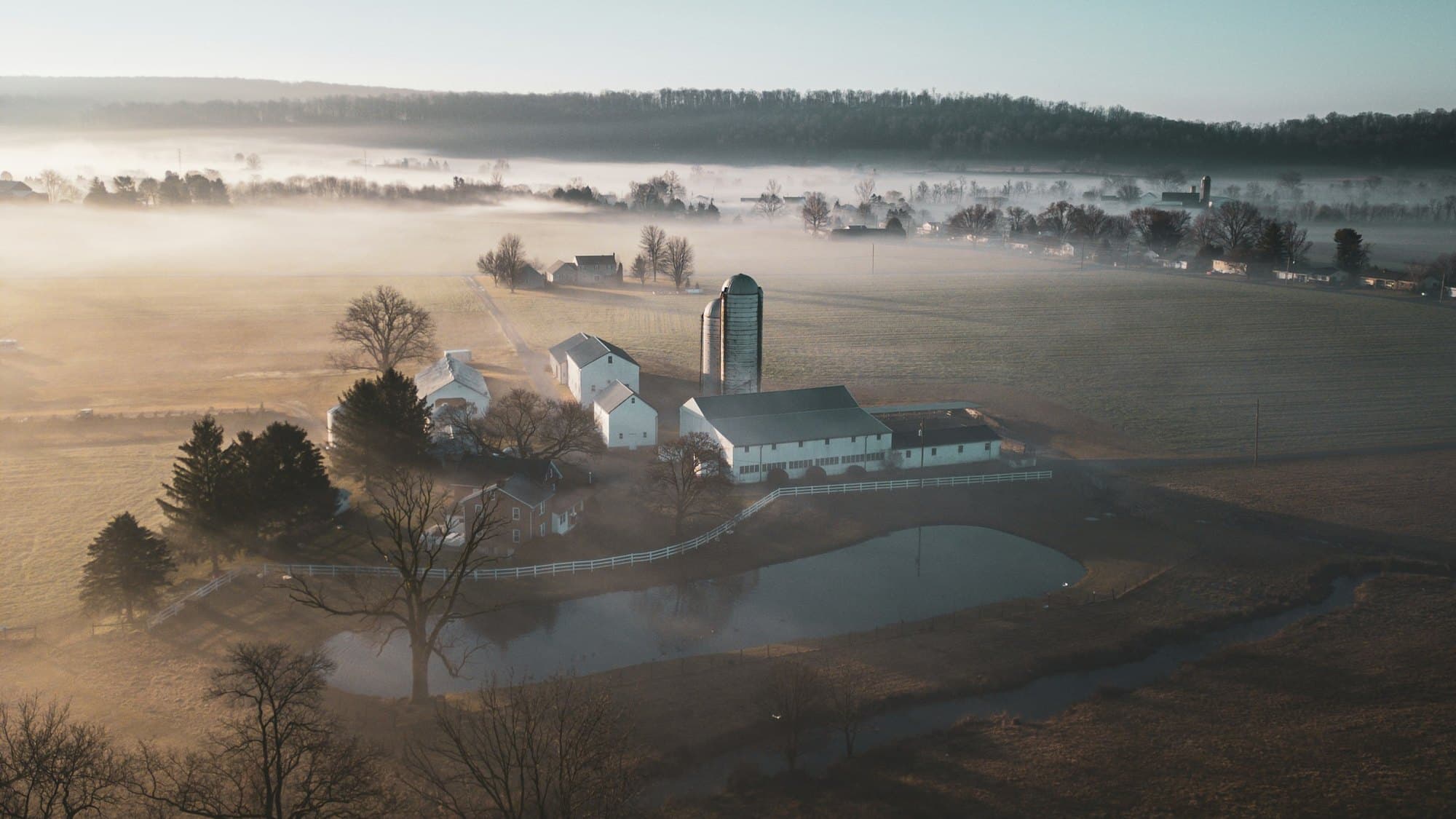 Aerial view of a farmstead at sunrise, white barn and two silos beside a pond in a misty valley